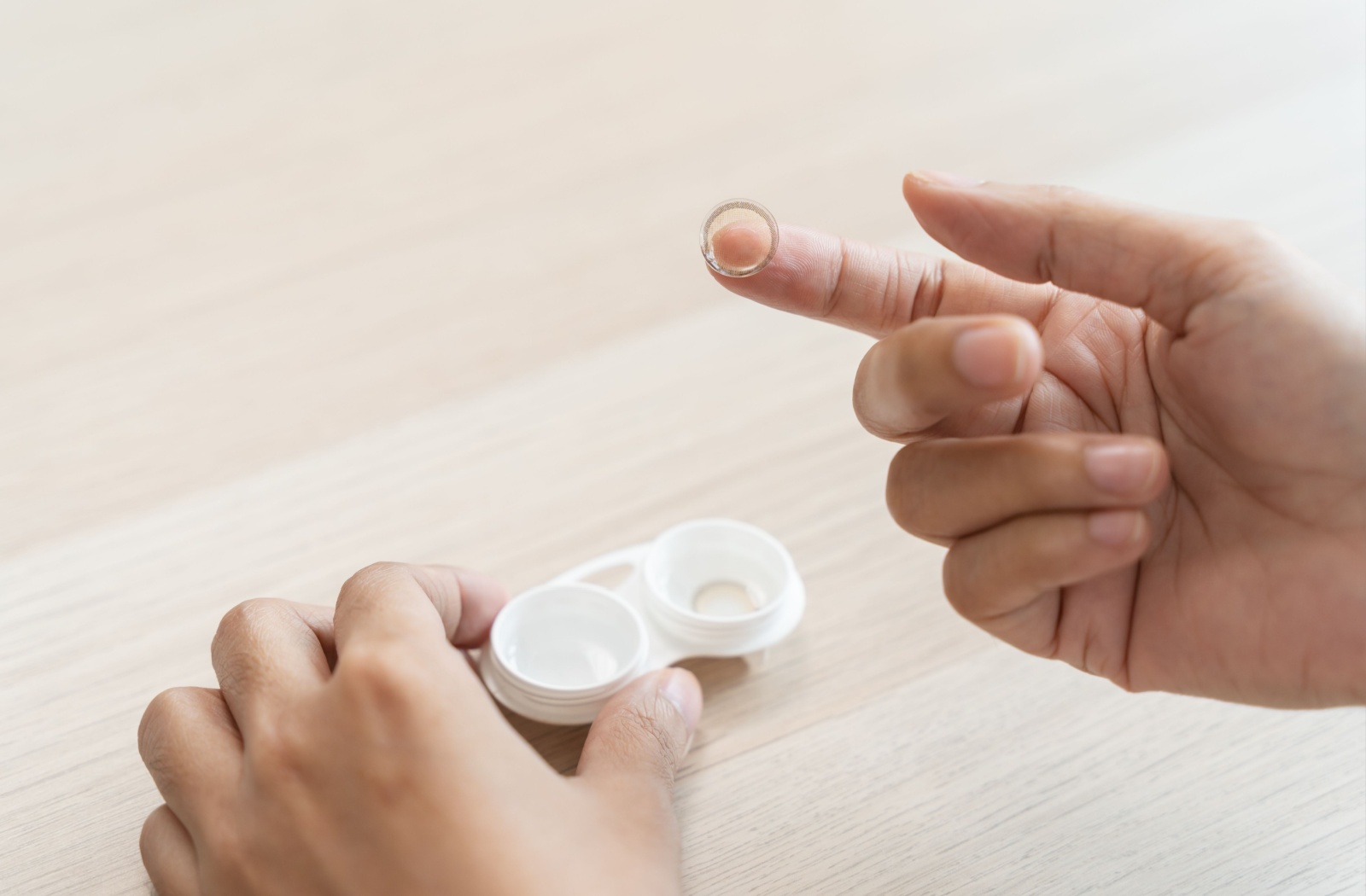 Close up of adult hands holding a contact lense on tip of finger, with contact lens case below.