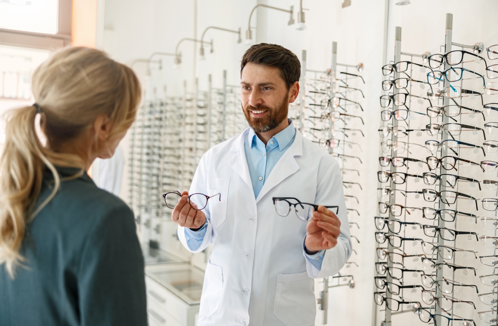 An eye care provider holds up two pairs of eyeglasses for their client to try on during a glasses styling appointment.