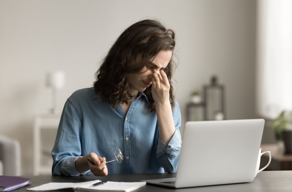 A person sits at a desk in front of a laptop, pinching the bridge of their nose with eyes closed in discomfort while holding their glasses in one hand.