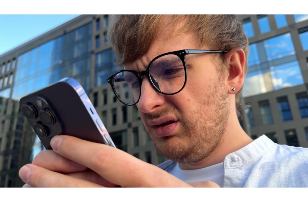 Man wearing glasses squinting at his smartphone outdoors, struggling to focus due to eye strain or vision issues.