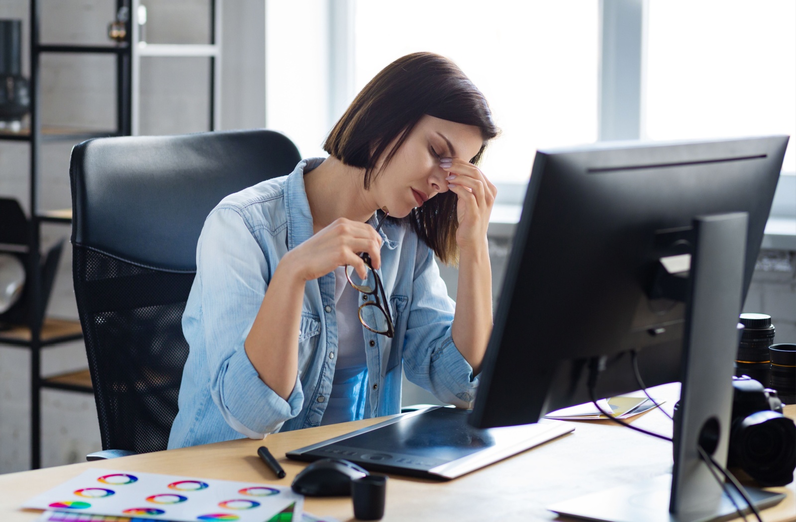 Woman sitting at her desk holding her glasses and rubbing her eyes from digital eye strain while working on a computer.