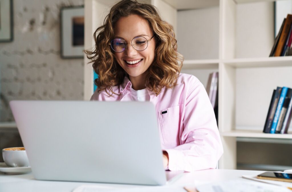 Woman wearing glasses smiling at her laptop in a bright office, showing comfortable and clear vision after reducing digital eye strain.