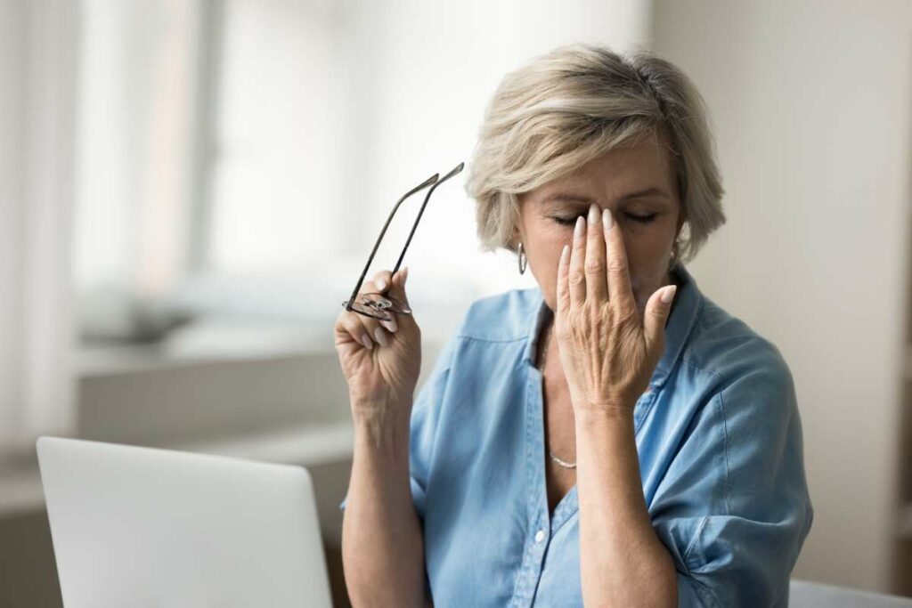 Older woman sitting at computer and rubbing her eyes while holding her glasses.